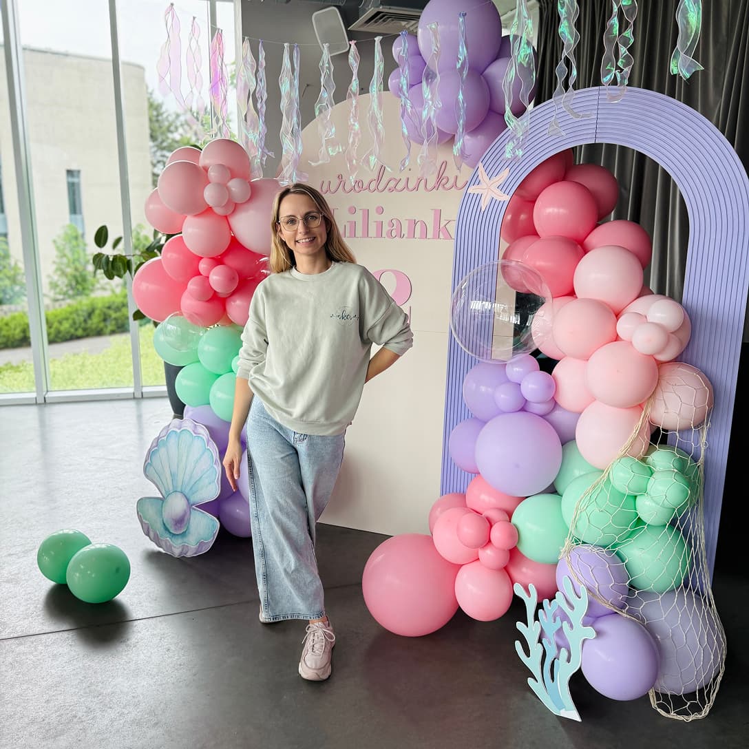 Uśmiechnięta dekoratorka przyjęć okolicznościowych pozująca przy kolorowej dekoracji w kształcie łuku z balonów, którą samodzielnie wykonała