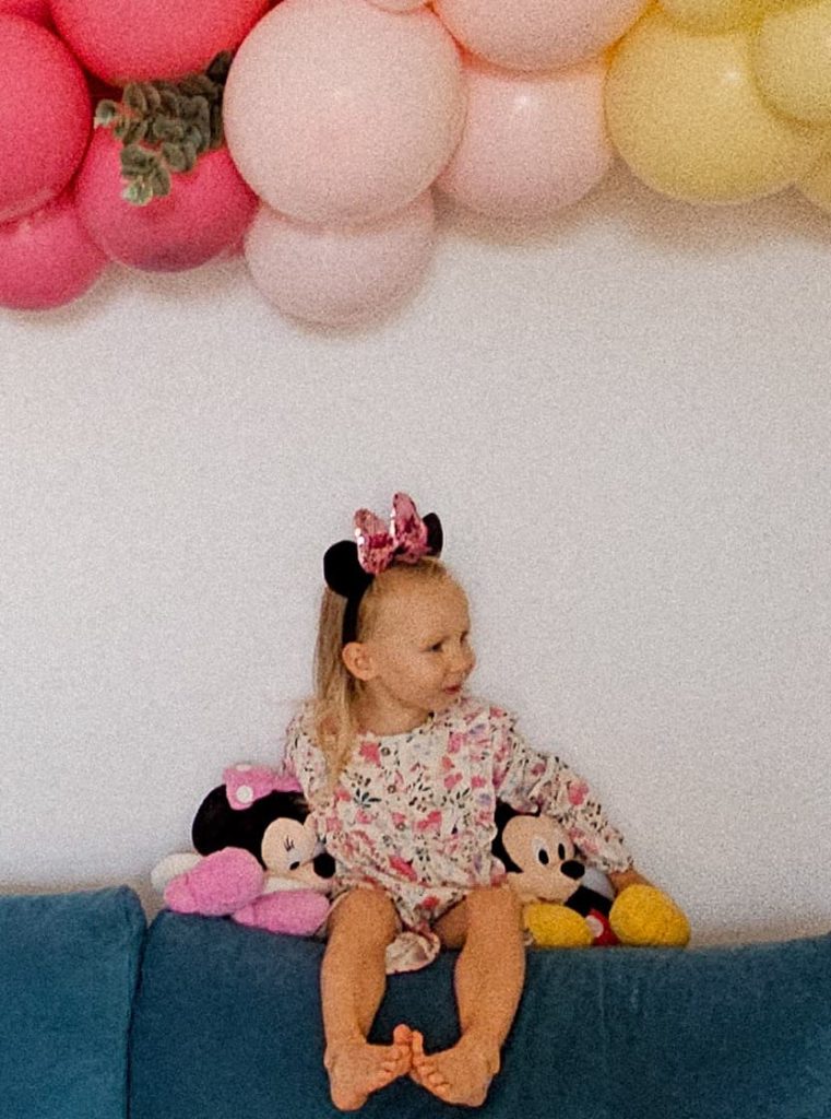 Mała dziewczynka w uszach Minnie na głowie siedząca na kanapie z pluszowymi zabawkami pod dekoracją z balonów