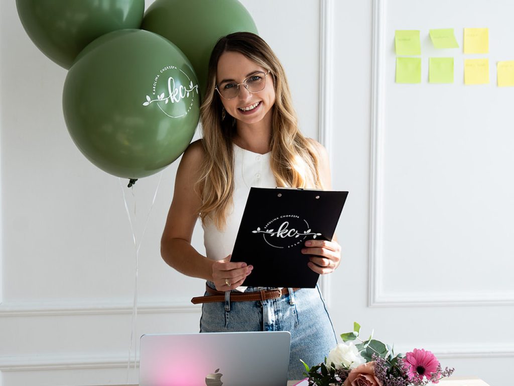 Uśmiechnięta Wedding Plannerka trzymająca podkładkę z klipsem i stojąca przed stolikiem na którym leży laptop i bukiet kwiatów. Obok wedding plannerki jest dekoracja balonów, a na ścianie przylepione są kolorowe karteczki samoprzylepne
