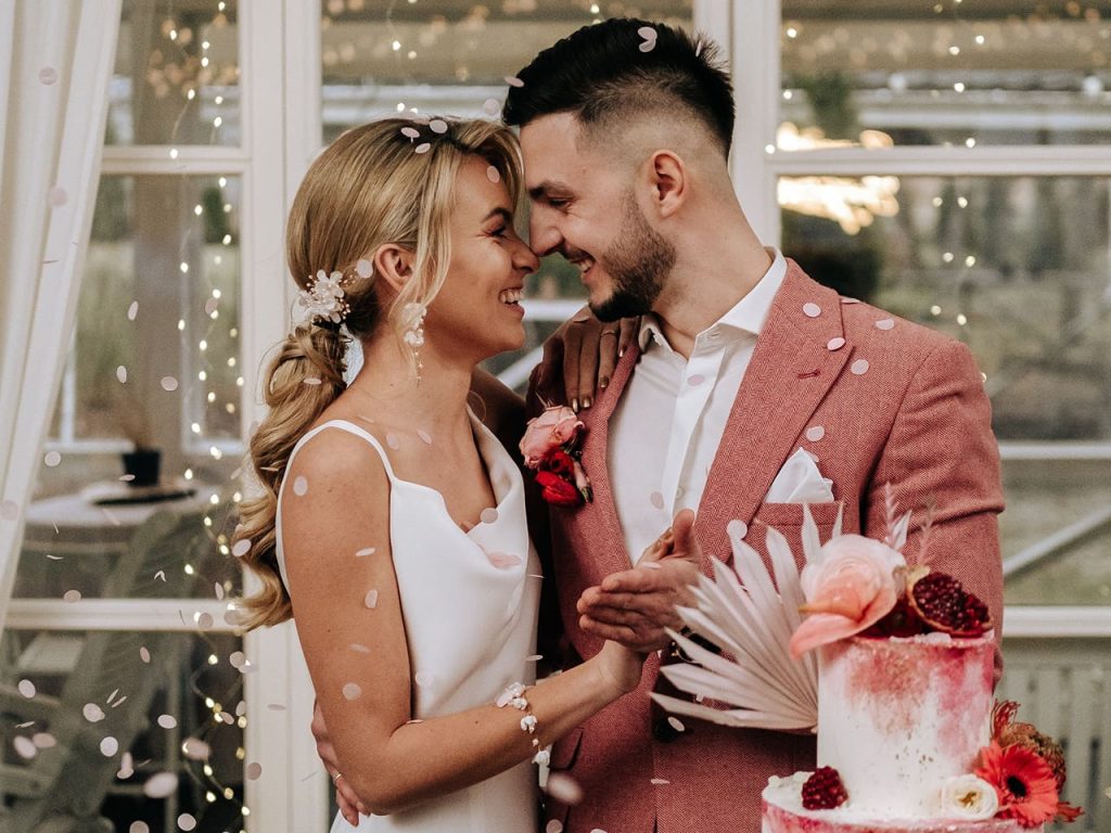 Uśmiechający się do siebie nowożeńcy stojący obok swojego tortu weselnego